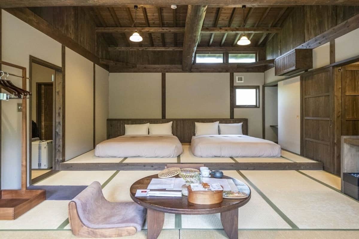 Quarto japonês tradicional com piso de tatame, duas camas, uma mesa de madeira com uma cadeira e teto com vigas aparentes.