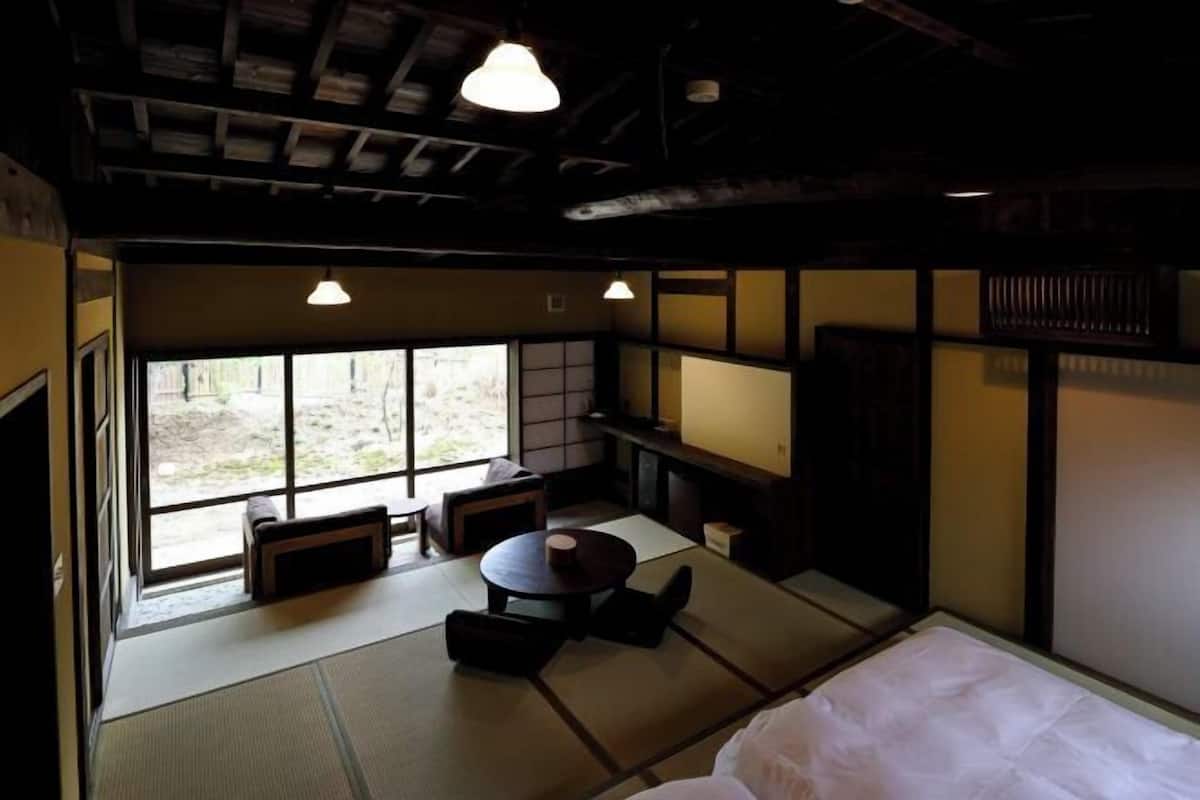 Um quarto japonês tradicional com tatames, uma mesa baixa, uma cama e grandes janelas.