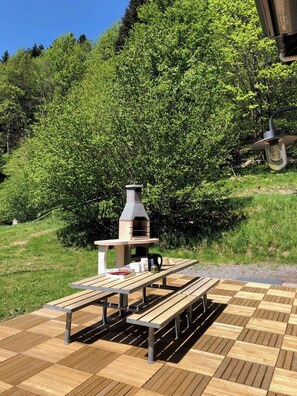 Um pátio externo com mesa e bancos de madeira, churrasqueira de pedra e um poste com luminária.
