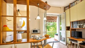 A kitchen with a wooden countertop, a dining area with a table and chairs, a television on a stand, and a balcony with a view of greenery.