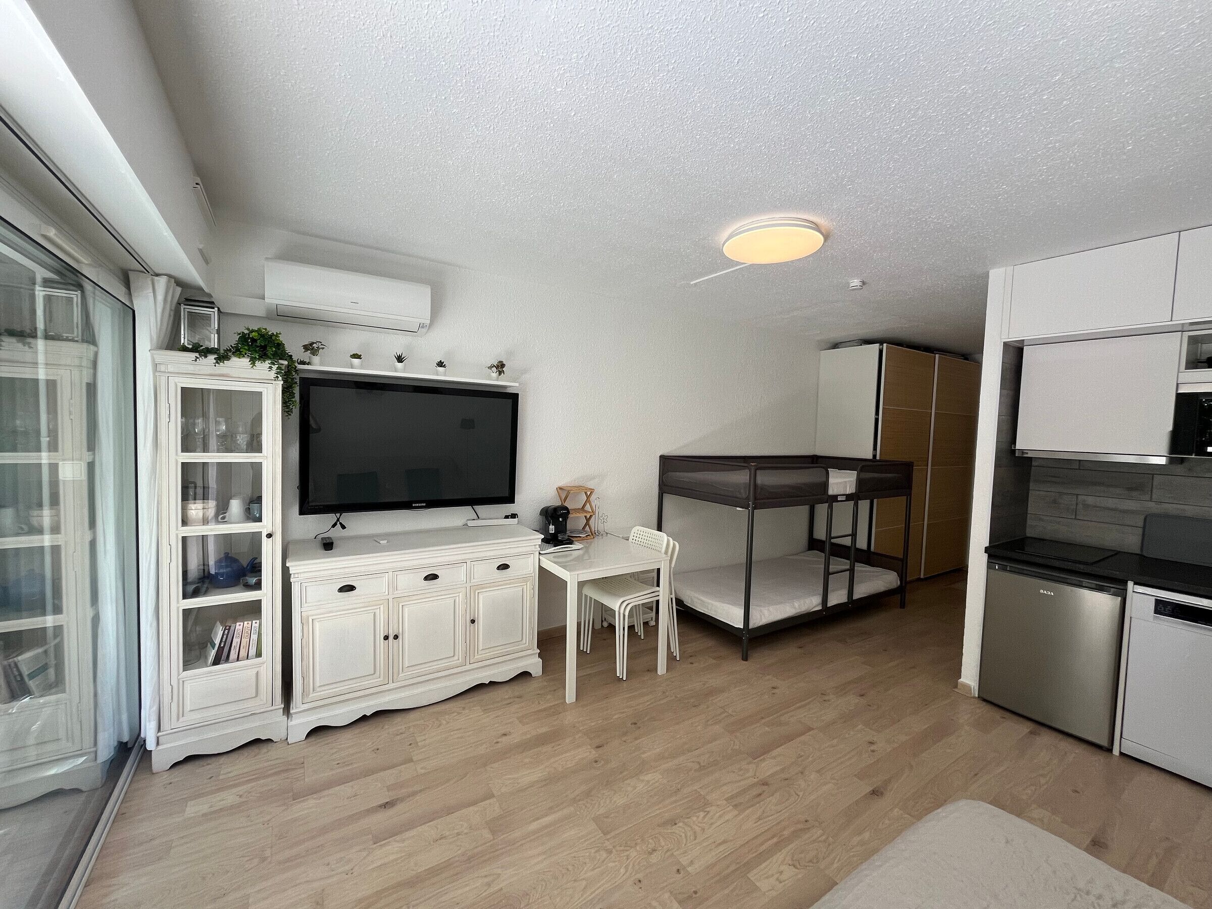 A compact kitchen with a built-in stove, a dining area with a white table and chairs, a bunk bed, and a TV on a cabinet.