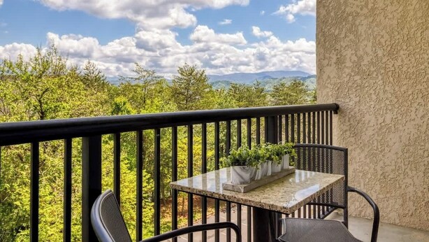 A balcony with a table and chairs, a potted plant, and a view of trees and mountains. - Mountain View Condo by Beyond Expectations: 2BR w/ Pools, Balcony & King Beds (Pigeon Forge)