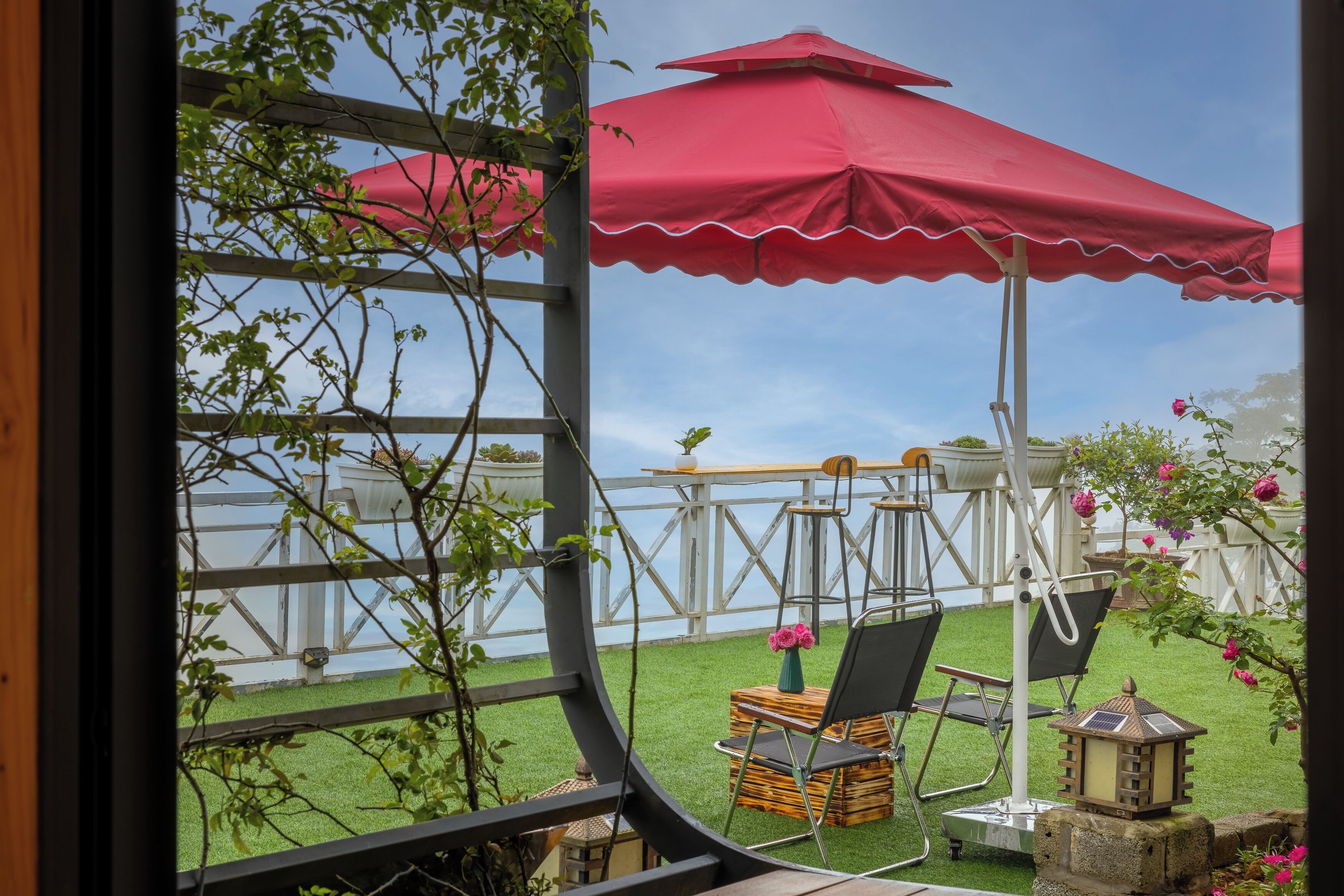 A view through a window of a patio area with a wooden deck, two red umbrellas, a table, and chairs.