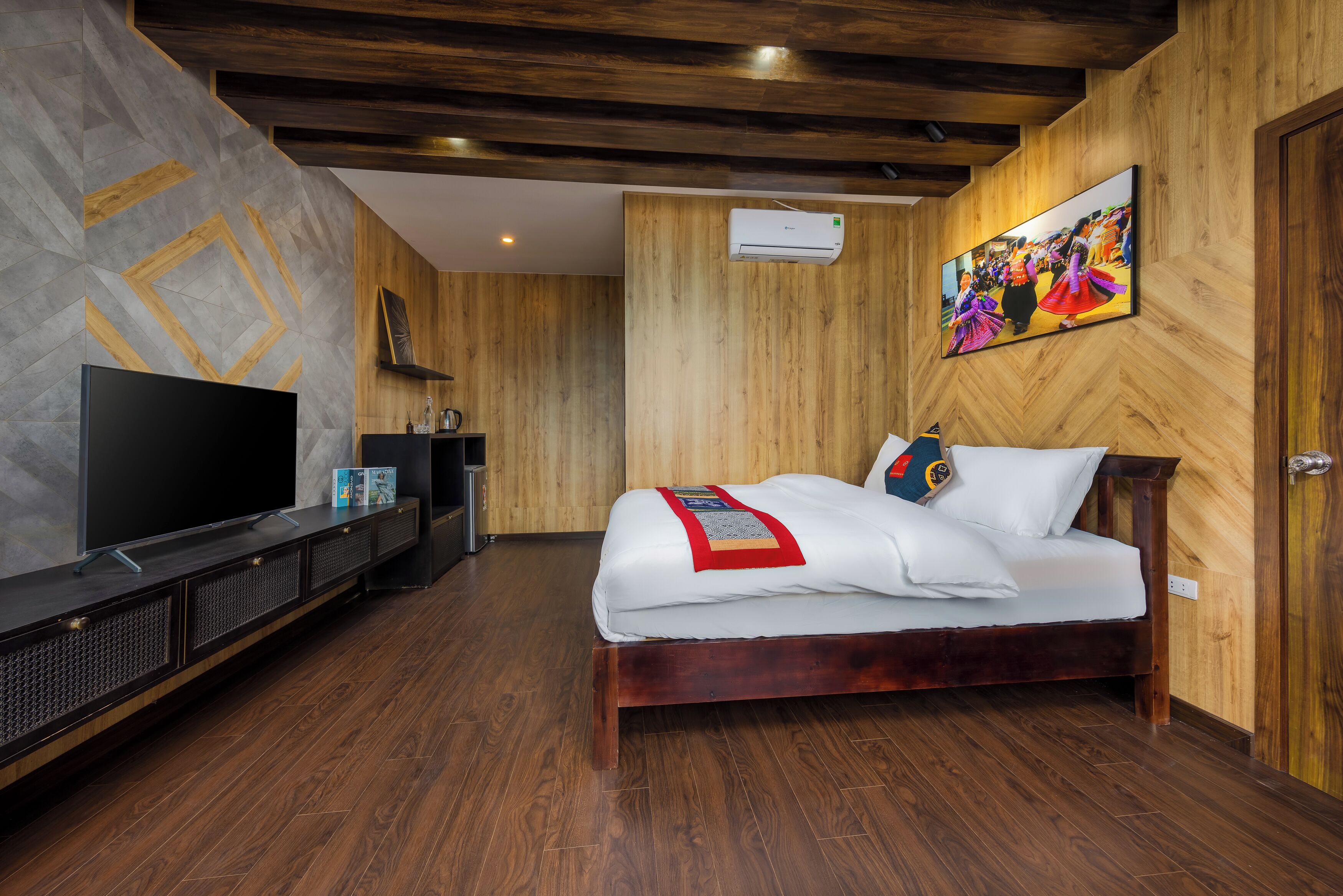 A modern hotel room with a wooden floor, a large flat-screen TV, a bed with white bedding, and a wooden headboard.