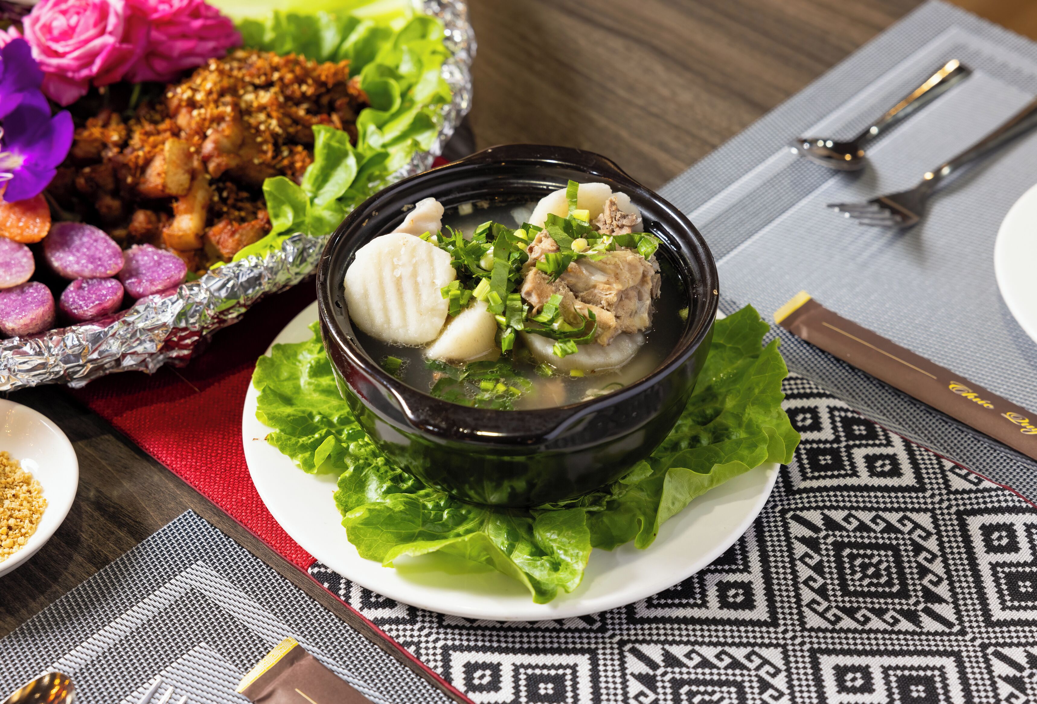 A bowl of soup with meat and vegetables on a plate with lettuce, accompanied by a side dish of fried items and purple flowers.