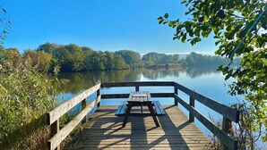 Ein hölzerner Steg mit Picknicktisch ragt in einen ruhigen See hinein, umgeben von Bäumen und Vegetation.