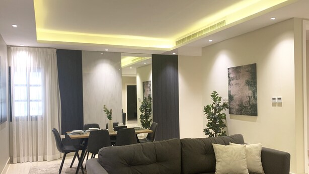 Ein modernes Wohnzimmer mit einer Couch, einem Esstisch und Stühlen. Ein großes Fenster mit Vorhängen, eine Pflanze und ein Bild an der Wand sind zu sehen.