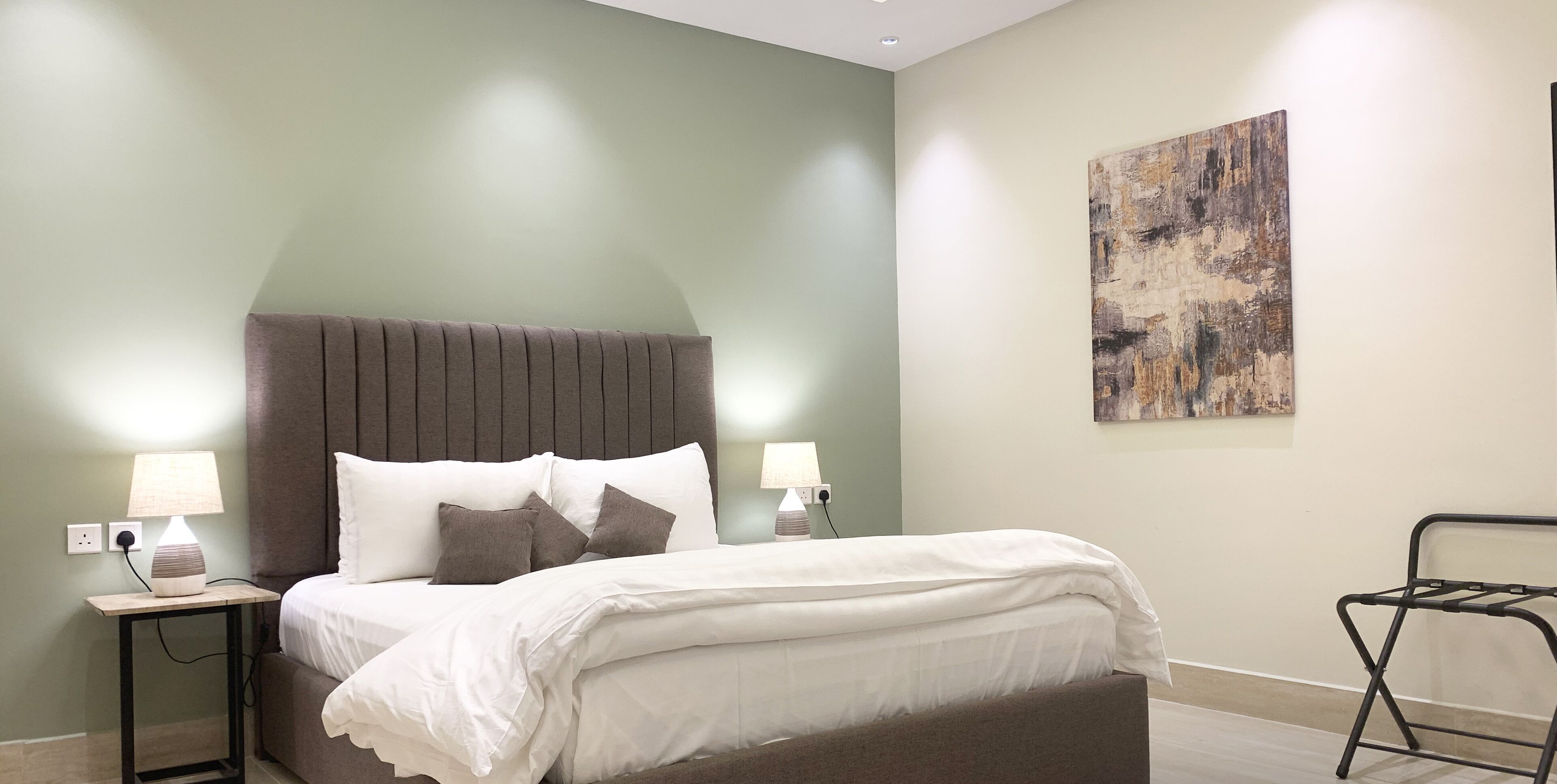 Une chambre à coucher comprenant un lit, deux lampes de chevet, une table de chevet, un tableau abstrait accroché au mur et une chaise en métal.