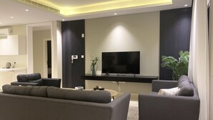 Ein modernes Wohnzimmer mit einem Flachbildfernseher, grauen Sofas, einem Couchtisch und einer Küchenzeile mit weißer Arbeitsplatte und Schränken.
