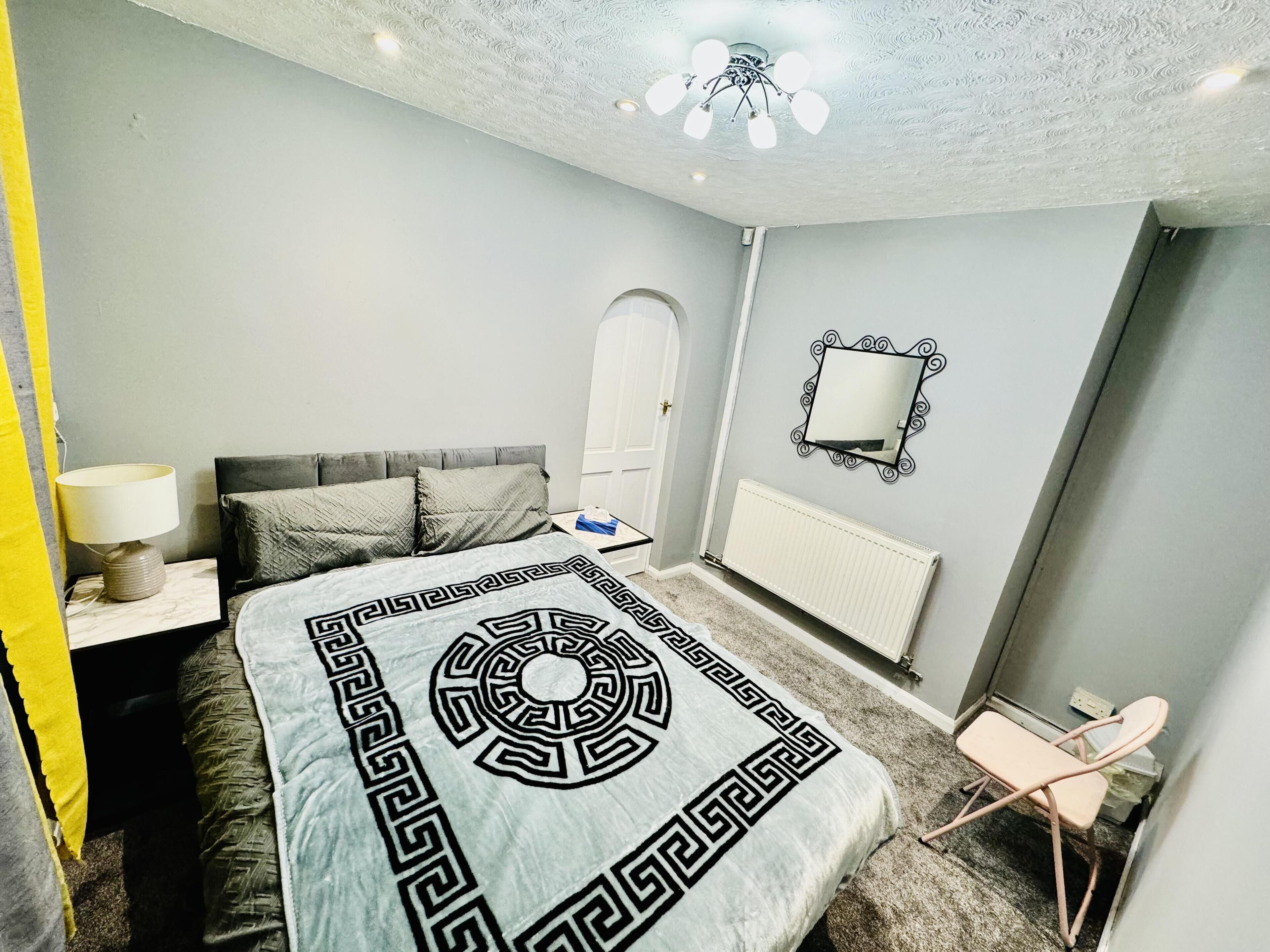 A bedroom with a bed, a nightstand, a chair, a radiator, a mirror, and a door.