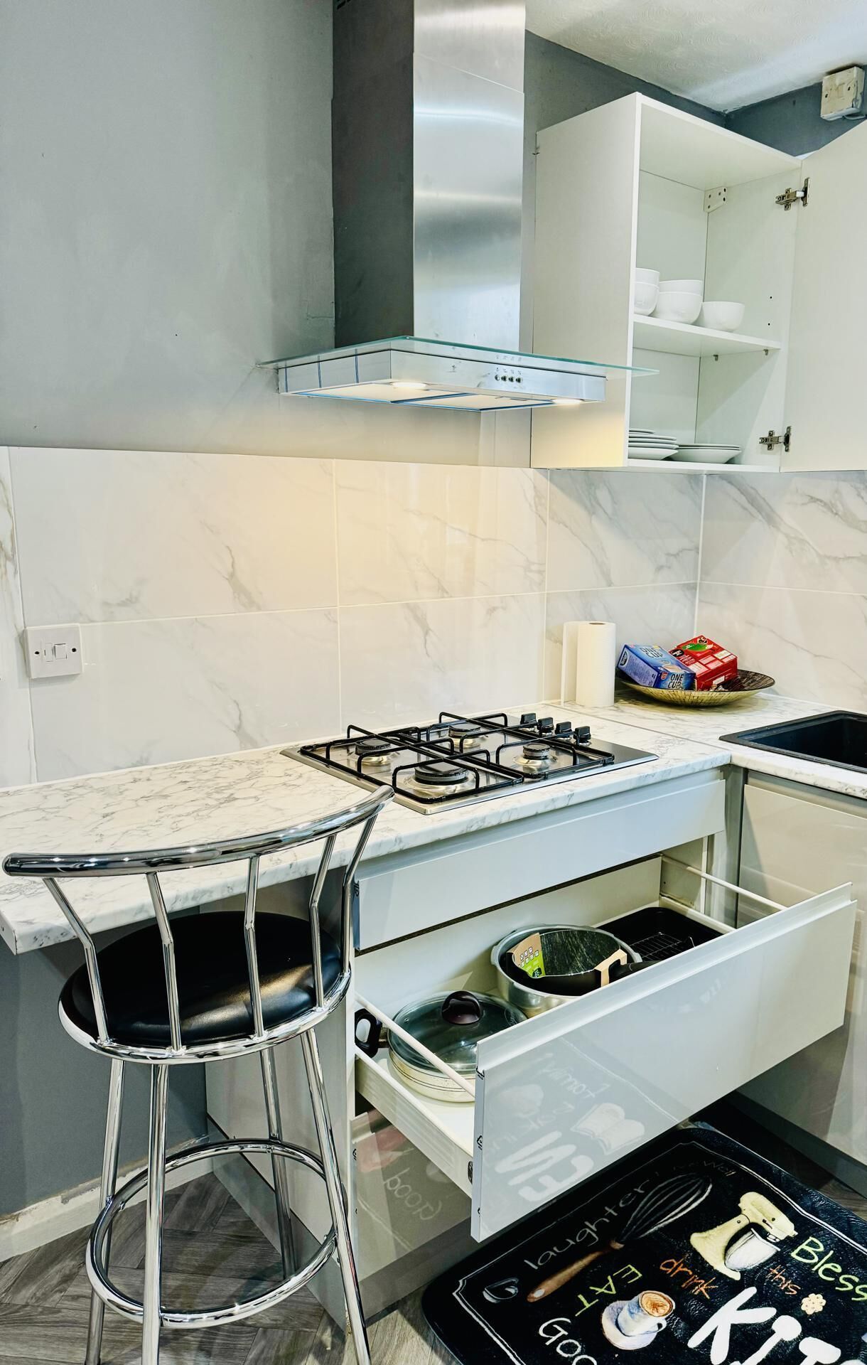 Una cucina moderna con piano cottura a gas, sgabelli da bar in acciaio inox, mobili bianchi e una parete a schiena di morsa in marmo.