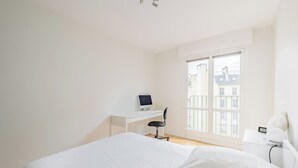 Un dormitorio con cama, escritorio con computadora, silla y ventana con vista a edificios.
