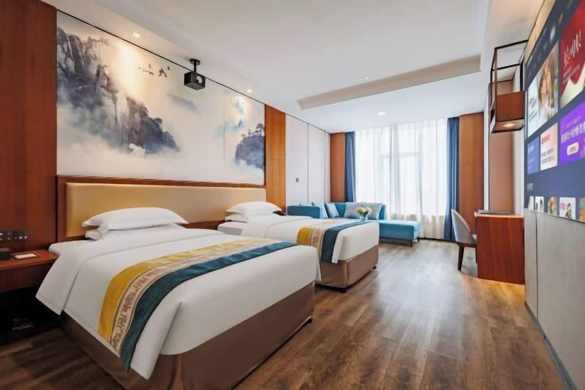 A hotel room with two beds, a desk, a chair, a large window with curtains, and a wall with artwork.