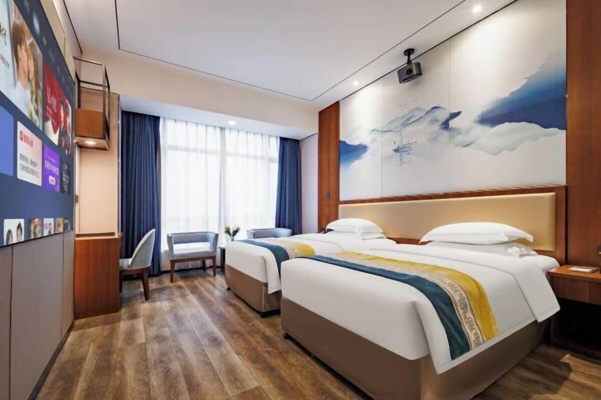 A hotel room with two beds, a desk, a chair, a large window with curtains, and a wall-mounted projector.