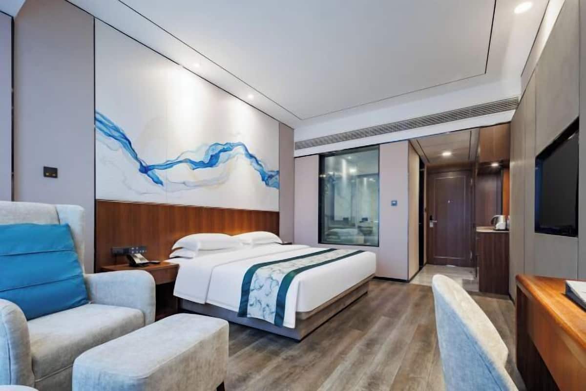 A modern hotel room with a large bed, a seating area, a TV, and a bathroom visible through a glass door.