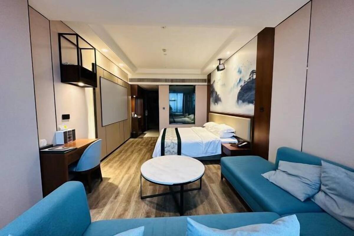 A hotel room with a bed, two blue sofas, a round table, a desk, and a large painting on the wall.
