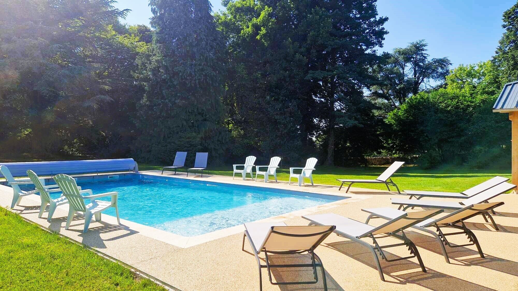 A rectangular swimming pool with a blue border, surrounded by a concrete deck. There are several white lounge chairs and a few Adirondack chairs arranged around the pool. The area is bordered by a well-maintained lawn and dense trees in the background.