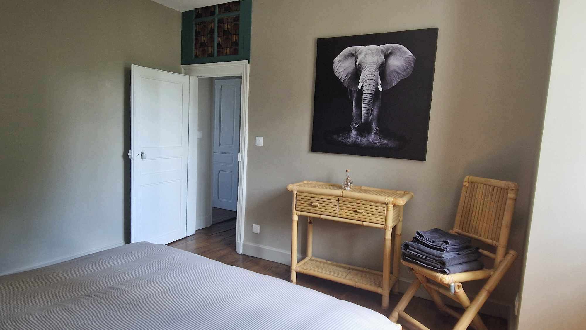 Un dormitorio con cama, mesita de noche, una silla, una puerta y un cuadro colgado en la pared con un elefante.