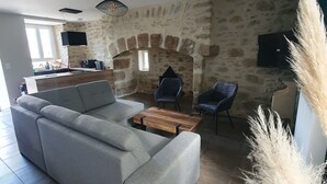 A modern living room with a grey sofa, a wooden coffee table, and two armchairs. A stone wall with a fireplace and a television mounted on it are visible. - Maison Rénovée Avec Terrasse + Parc Avec Piscine Chauffée et Terrasse Couverte (Estivaux)
