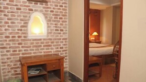 A room with a brick wall, a wooden nightstand with a lamp, a bed, and a chair.