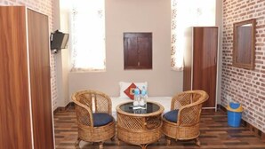 A room with a brick wall, a bed with a pillow, two wicker chairs, a small table with bottles, and a TV mounted on the wall.