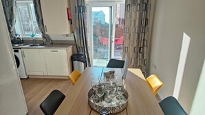 Zona pranzo con tavolo di legno apparecchiato per sei, cucina sullo sfondo e vista su un edificio rosso attraverso una porta vetrata.
