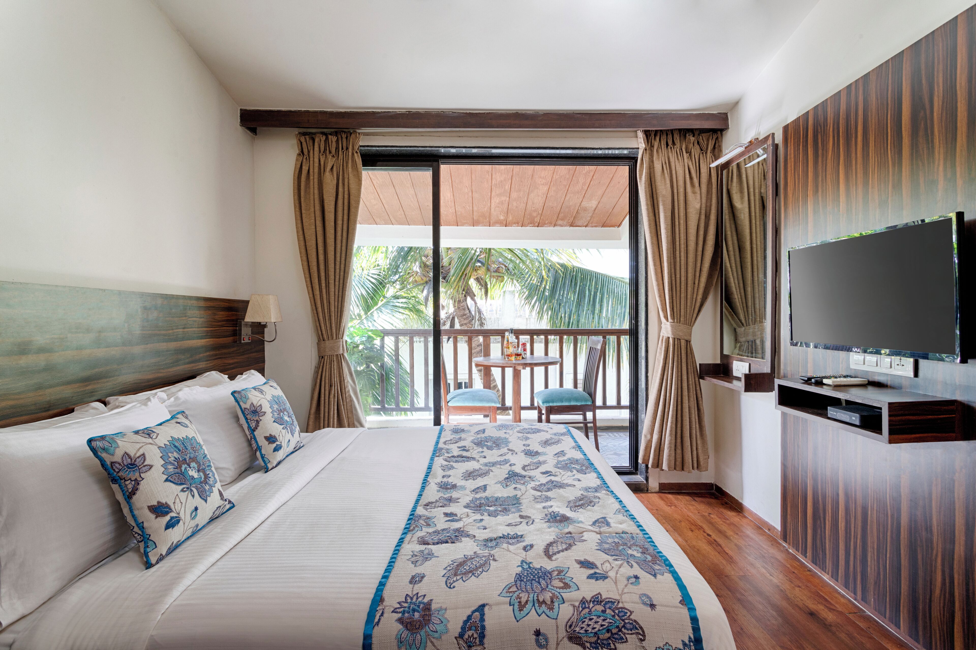Habitación de hotel con una cama grande, un televisor montado en la pared, un balcón con mesa y sillas, y vista a palmeras.