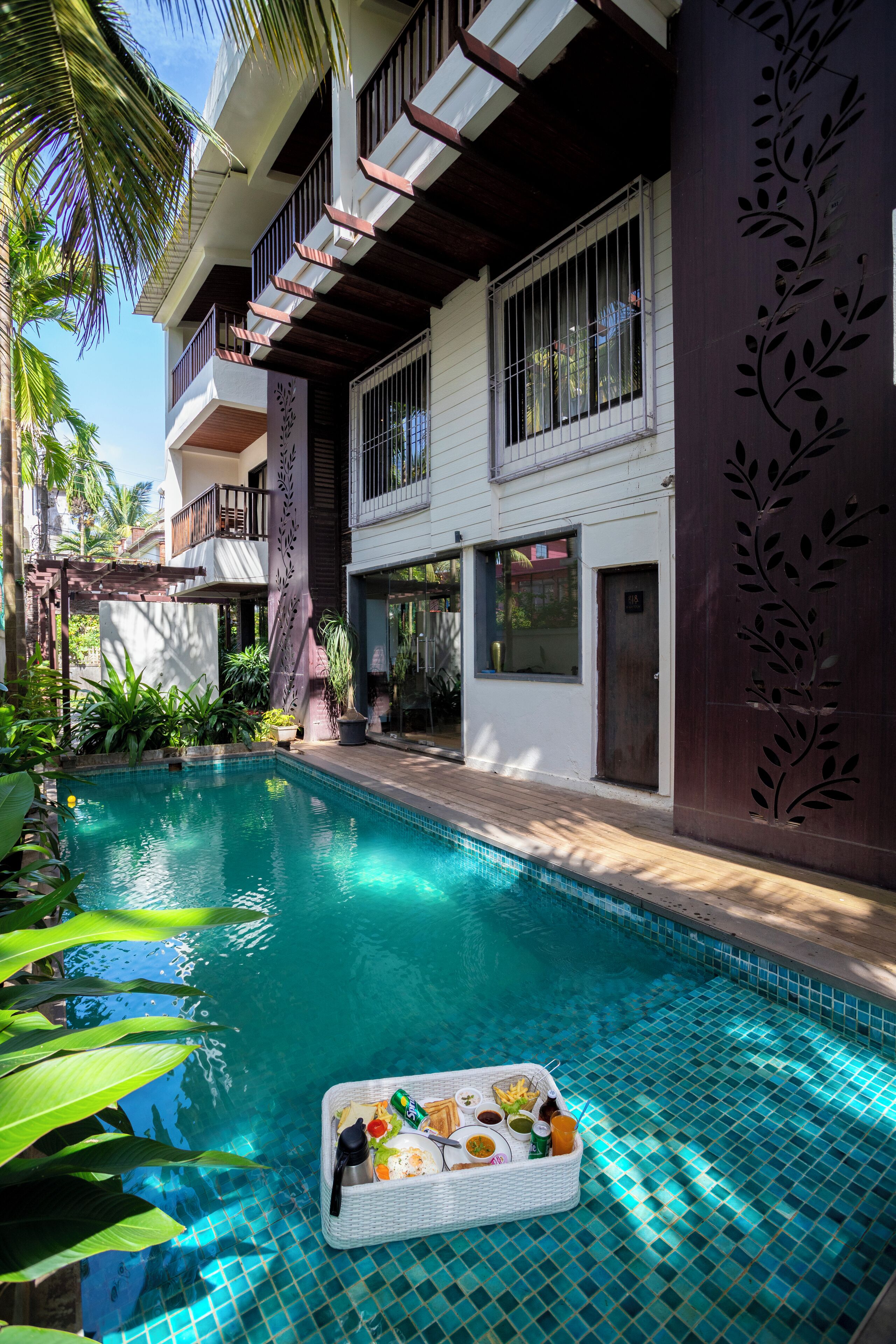 A swimming pool with a floating tray of food, surrounded by lush greenery and a multi-story building with balconies.