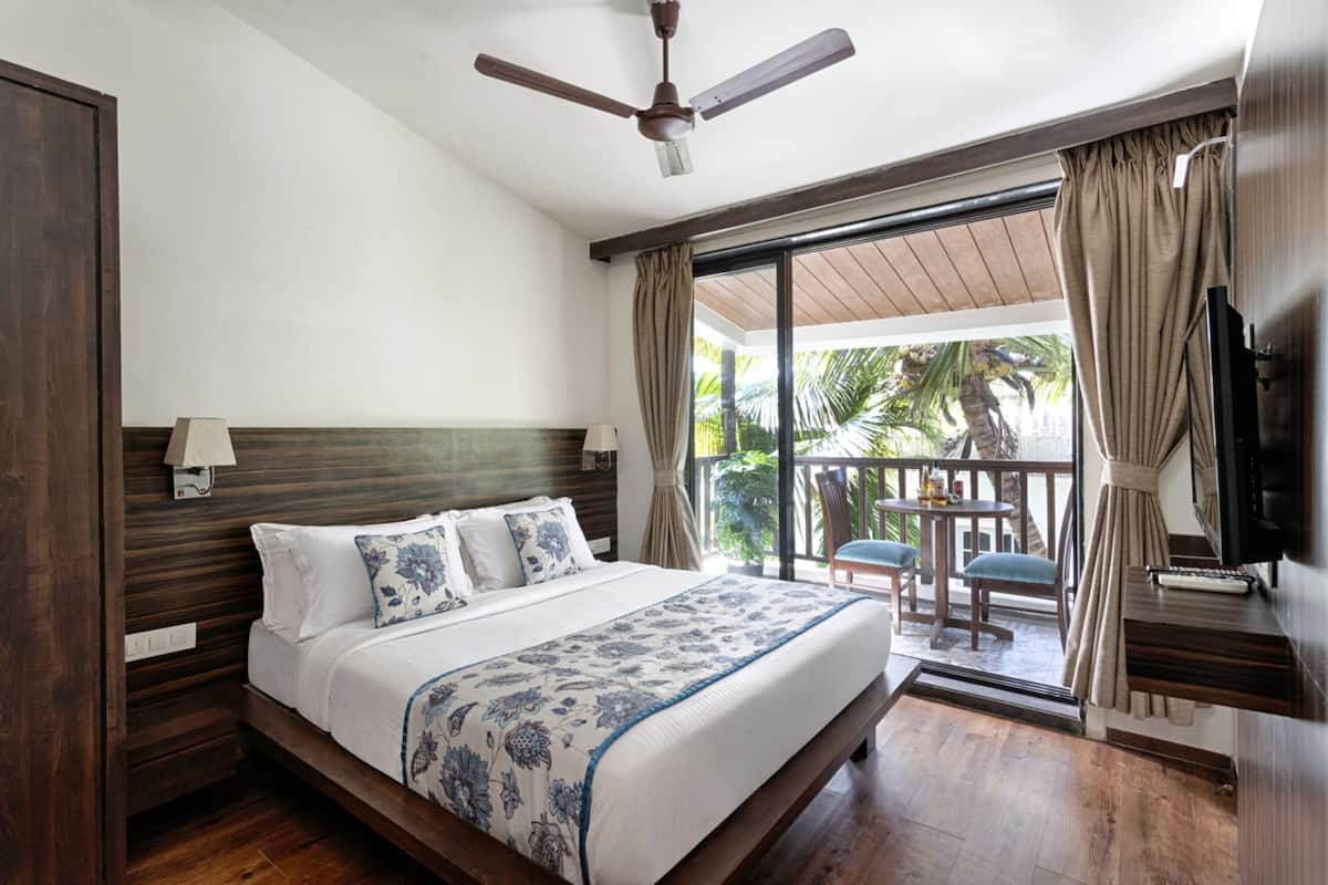 A hotel room with a large bed, a ceiling fan, a balcony with a table and chairs, and a TV on the wall.