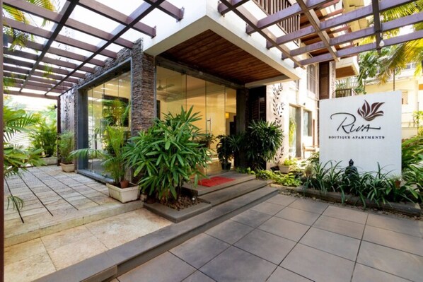 A modern building with a glass entrance, a wooden pergola, and a sign for 'Riva Boutique Apartments'. - Riva Boutique Apartments (Alto de Porvorim)