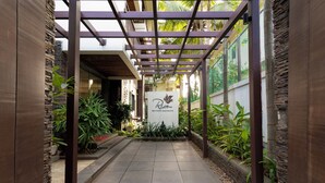 A covered walkway with a stone wall on one side and a metal fence on the other, leading to a building with a sign that reads "Ria Boutique Apartments." - Riva Boutique Apartments (Alto de Porvorim)