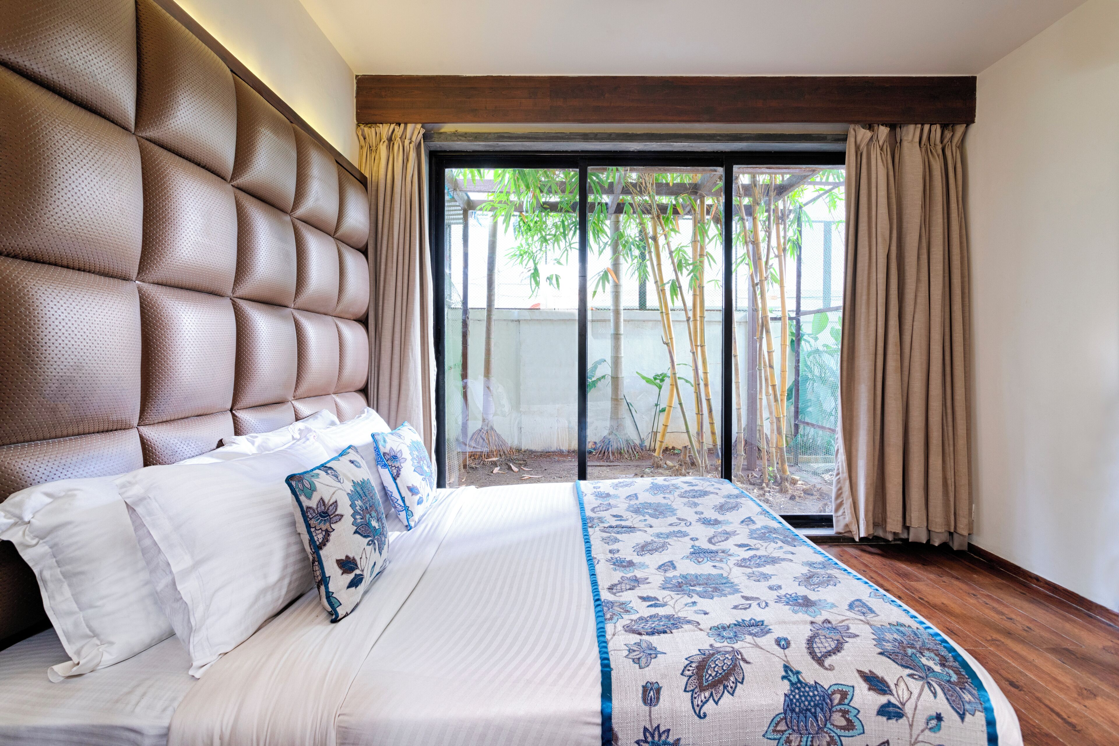 Un dormitorio con cabecera capitonada, una cama grande con ropa de cama blanca y azul estampada con motivos florales, y vista al bambú a través de una puerta corrediza de vidrio.