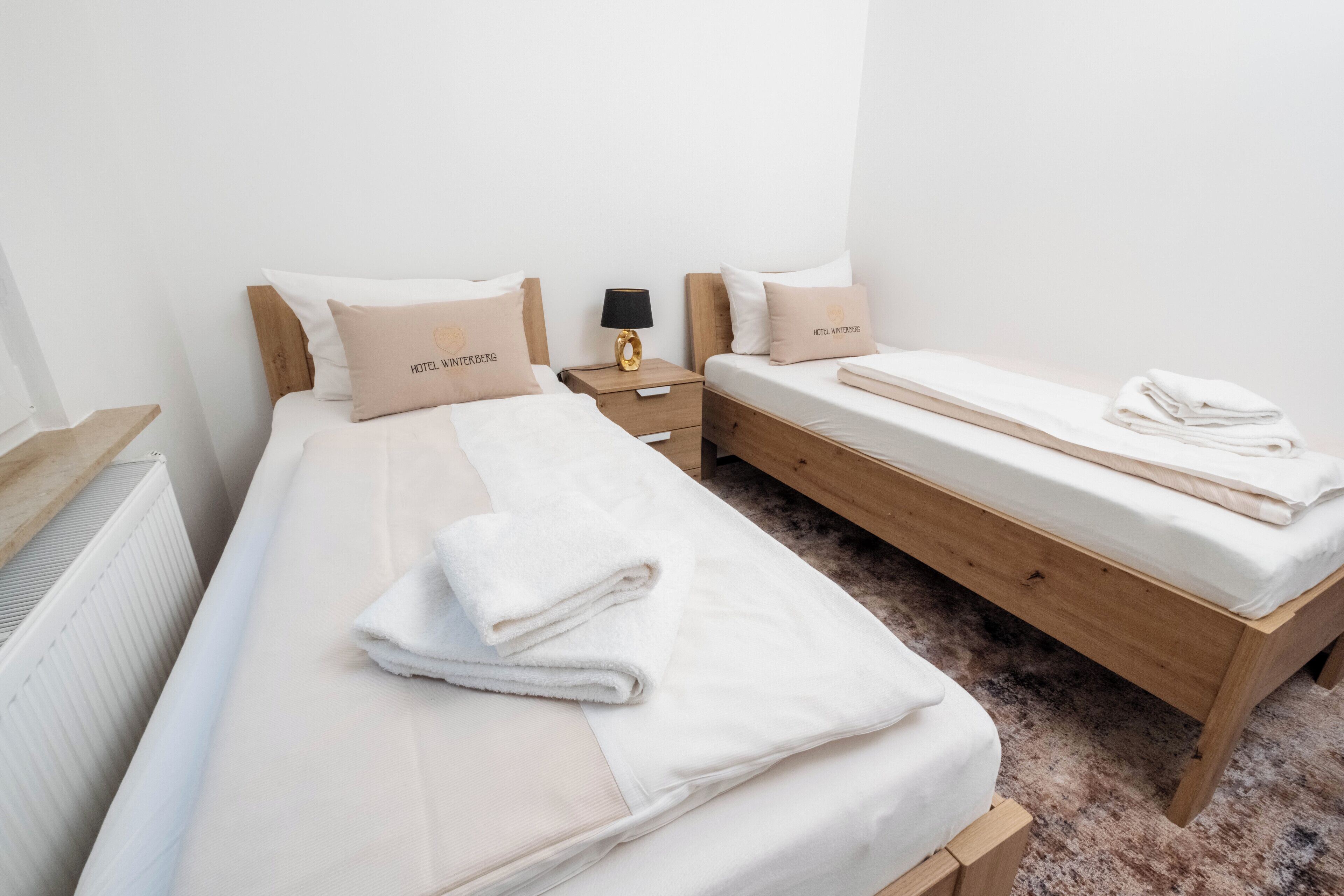 Habitación de hotel con dos camas, una mesita de noche de madera, un radiador y una pequeña mesa con una lámpara.