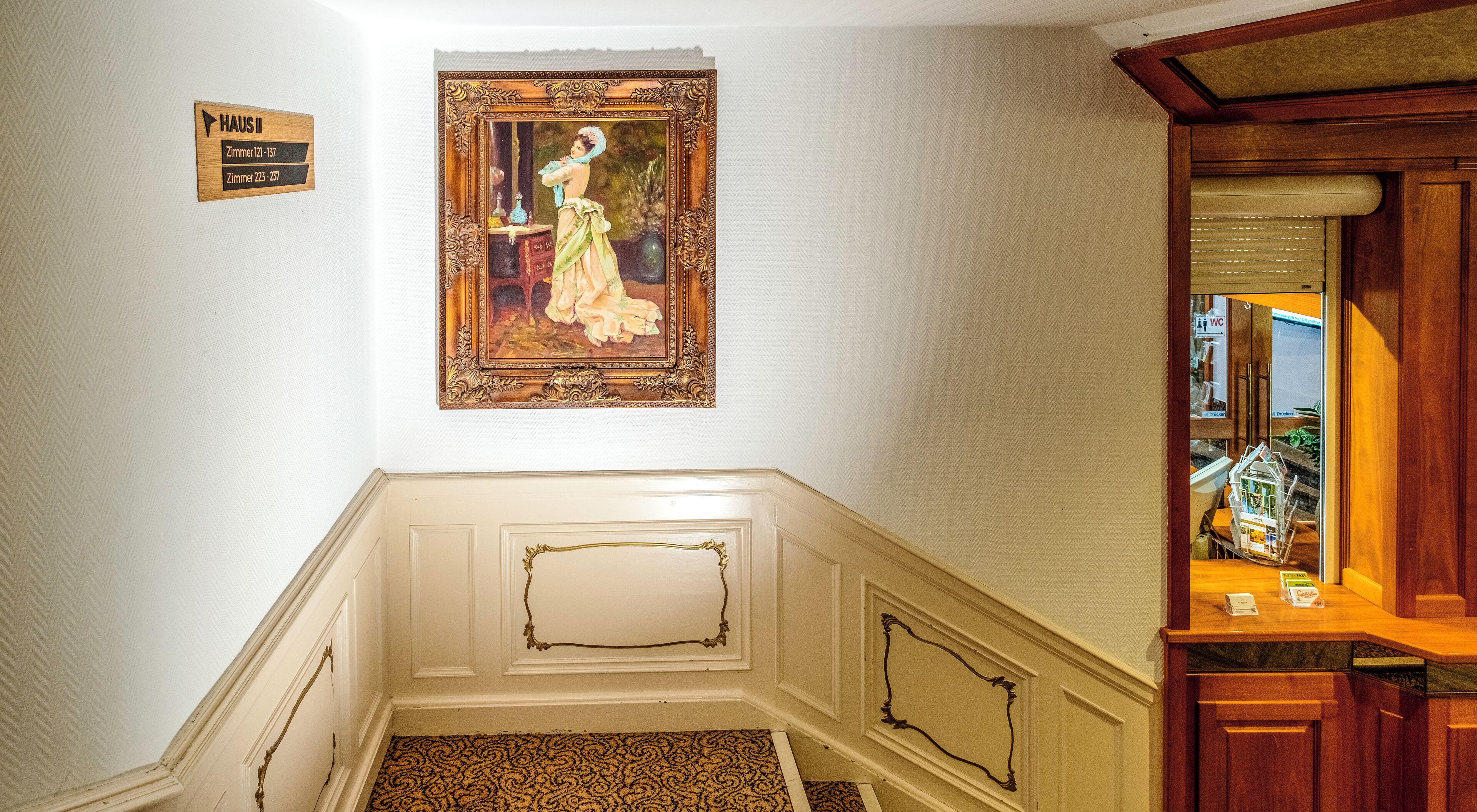 A hallway with a wooden sign indicating room numbers, a framed painting on the wall, and a wooden counter with items on it.