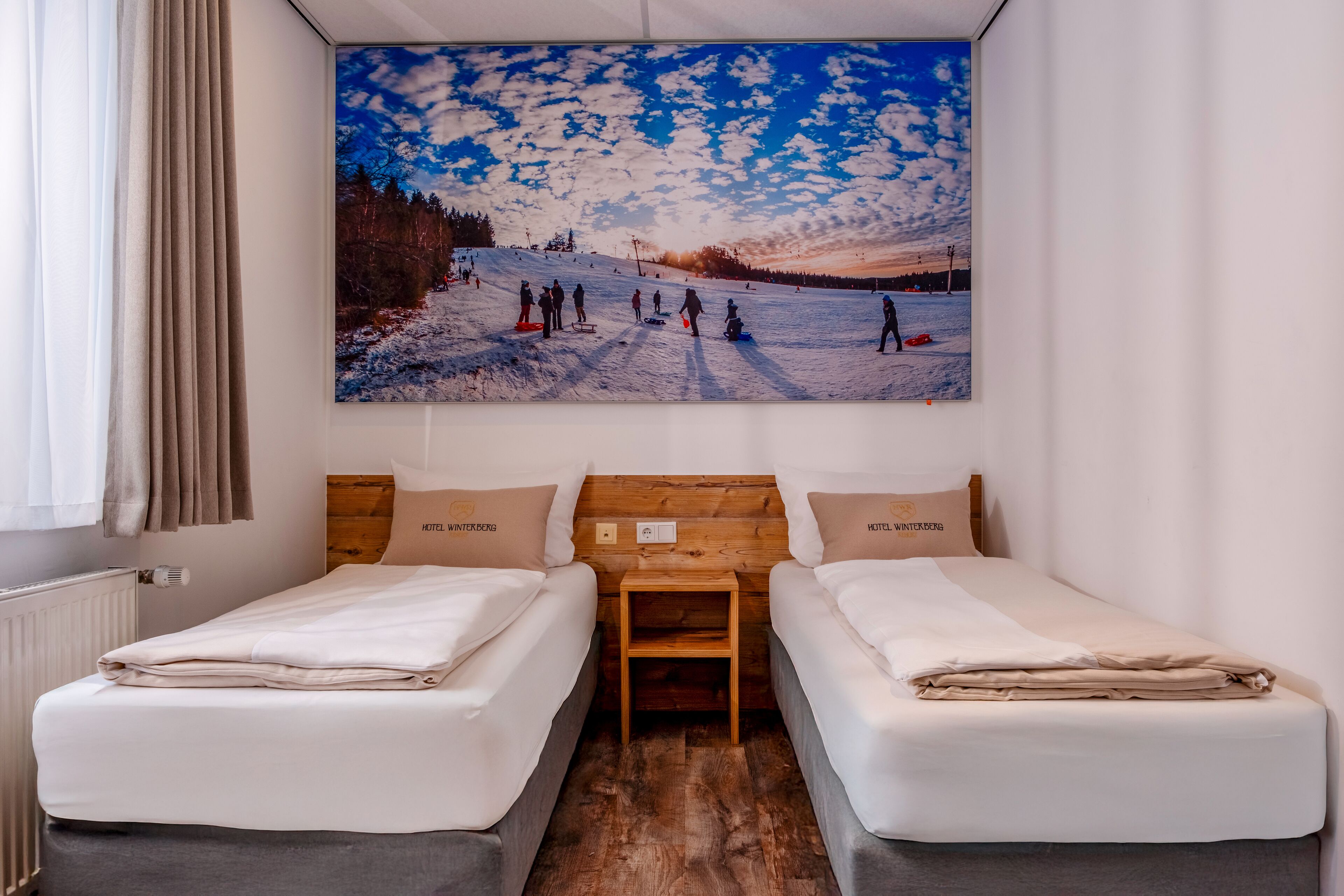 A hotel room with two beds, a wooden headboard, a painting of a snowy landscape, and a window with curtains.