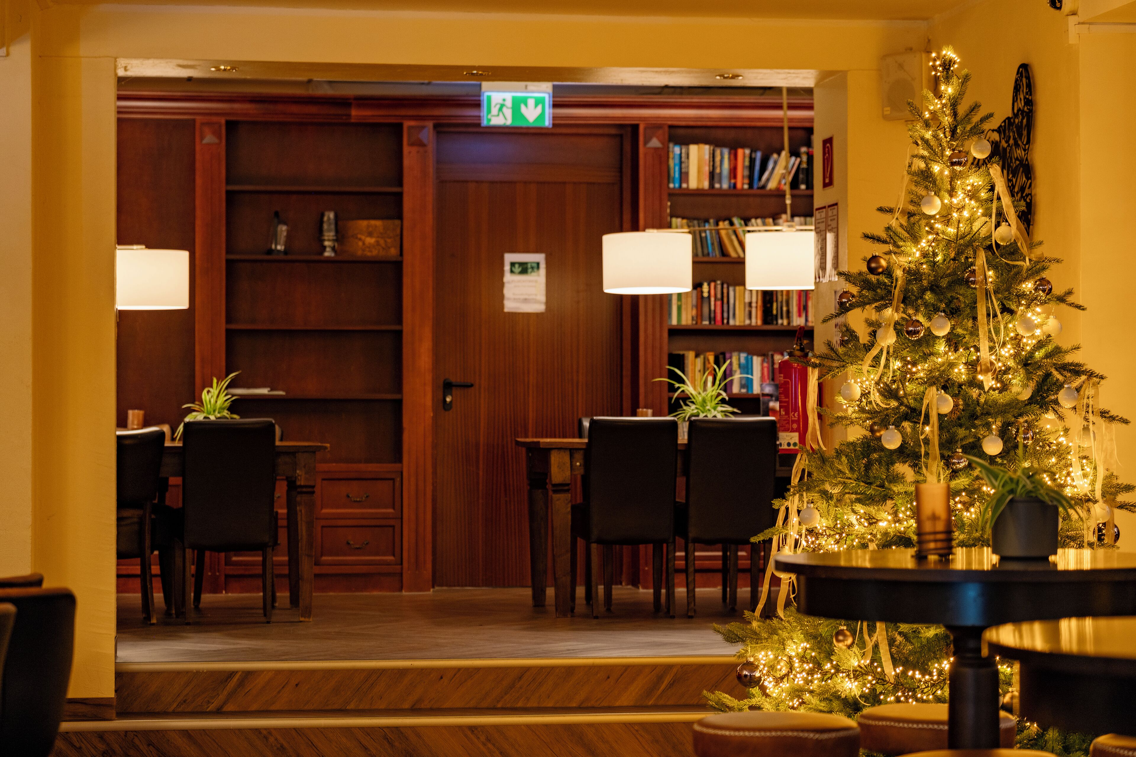 Een sfeervol verlichte kamer met een kerstboom, een boekenkast en een eethoek met stoelen en tafels.