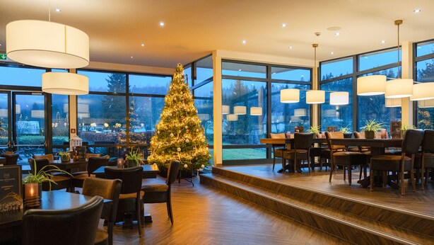 A modern dining area with a Christmas tree, wooden flooring, and large windows offering a view of the outdoors. - Hotel Winterberg Resort (Winterberg)