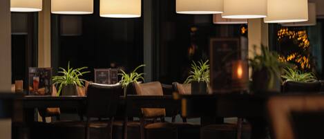 A dimly lit restaurant interior with multiple tables and chairs, potted plants, and hanging lamps.