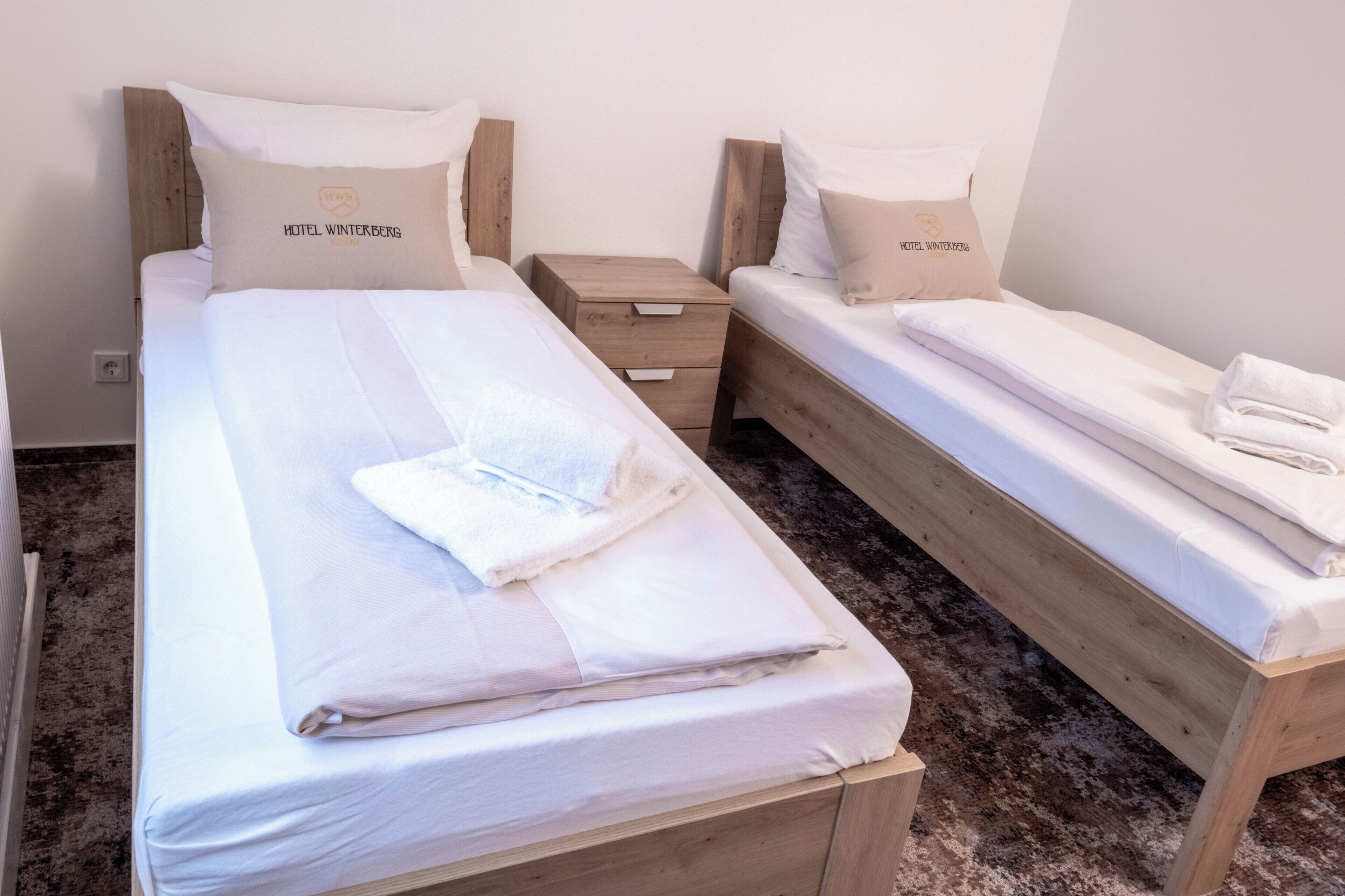 A hotel room with two single beds, each with a wooden headboard and matching pillows. There is a small wooden nightstand between the beds with a drawer.
