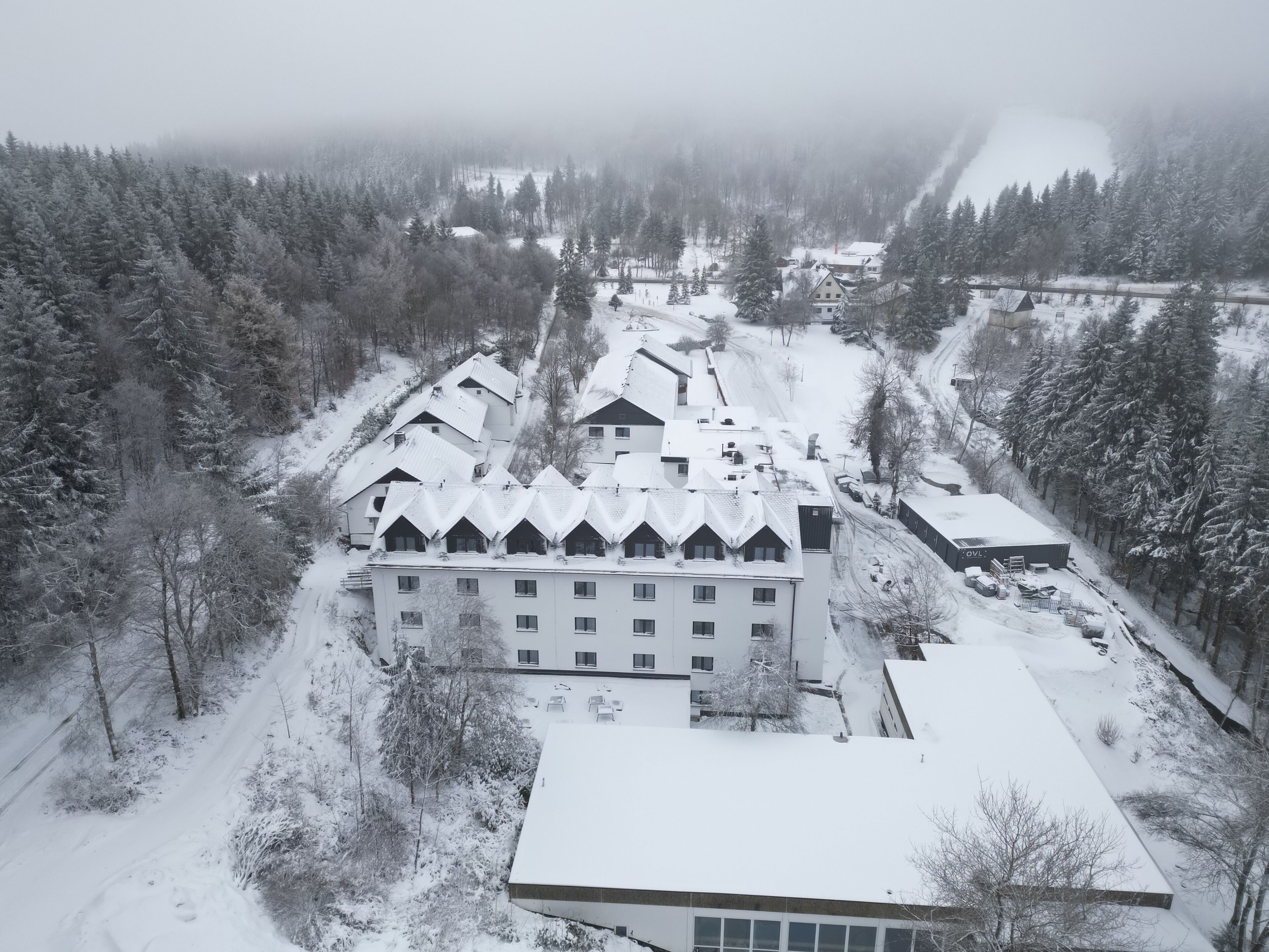 A large, multi-story building with a symmetrical roofline, surrounded by snow-covered trees and a forested area.