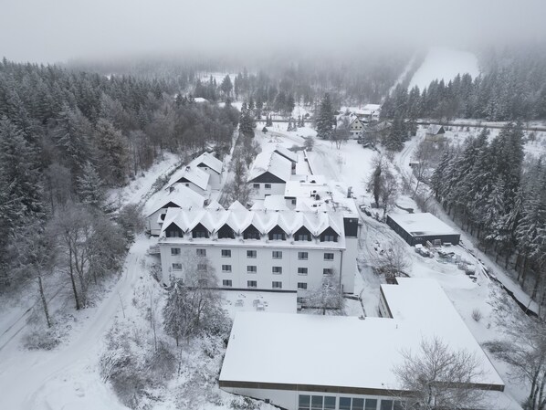 A large, multi-story building with a symmetrical roofline, surrounded by snow-covered trees and a forested area.
