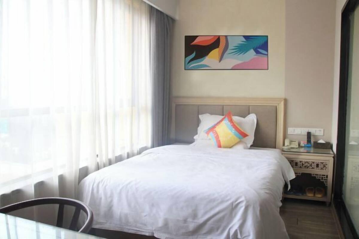 A hotel room with a bed, a bedside table, a chair, a window with curtains, and a painting on the wall.