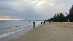 Uma praia com pessoas caminhando pela orla, areia dourada e céu nublado.