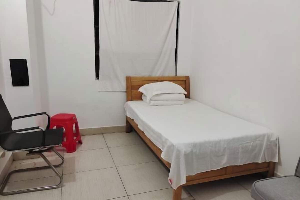 A small, clean room with a single bed, a chair, and a red stool.