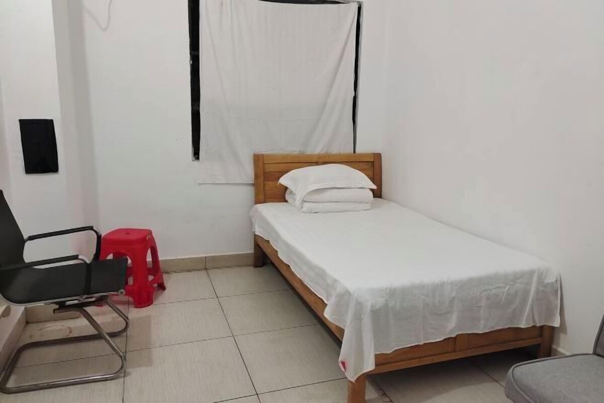 Quarto pequeno e limpo com uma cama de solteiro, uma cadeira e um banquinho vermelho.