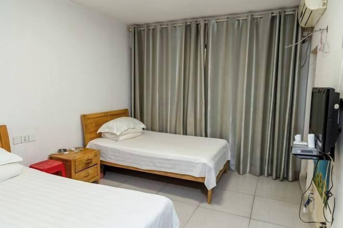 A hotel room with two beds, a wooden headboard, a red stool, a TV, and a curtain.
