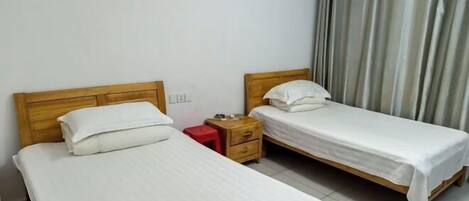 Une petite chambre propre, avec deux lits simples, une table de chevet en bois et un tabouret rouge.