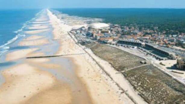 An aerial view of a coastal area with a long sandy beach, a dense urban area with buildings, and a clear blue sky. - Maison 130 m2 sur Jardin 600 m2 à 900 m de la Plage et Centre Ville-4 Chambres (Lacanau)