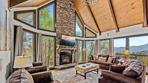 A spacious living room with a stone fireplace, leather sofas, and large windows offering mountain views.