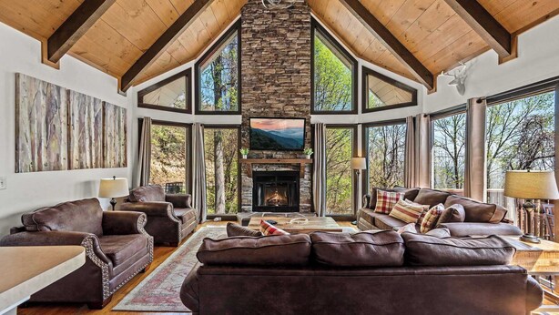 A spacious living room with a stone fireplace, leather sofas, and large windows offering a view of the outdoors.